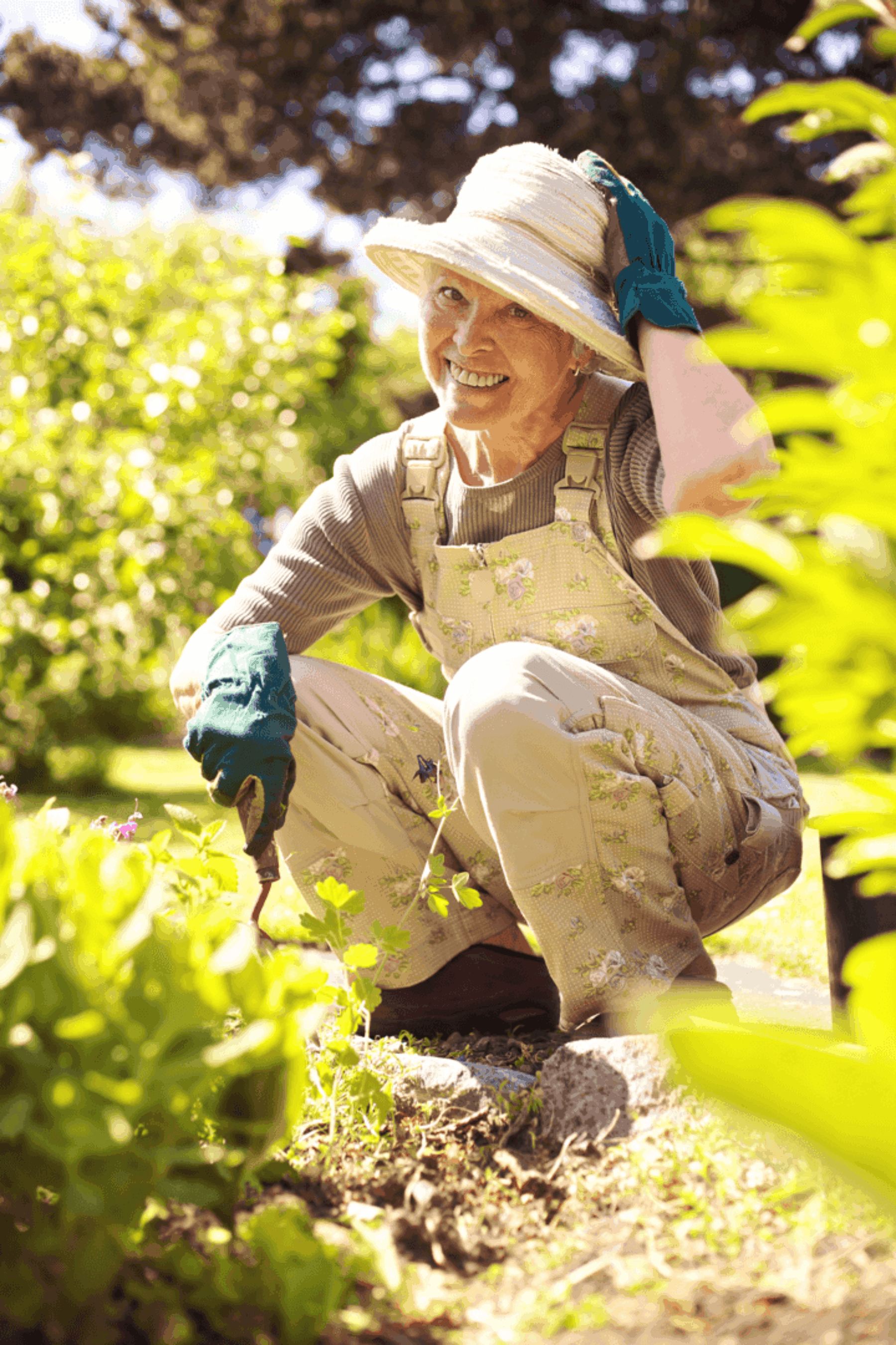 Woman squatting down while gardening, surrounded by plants, has her hand on her wide-brimmed hat.