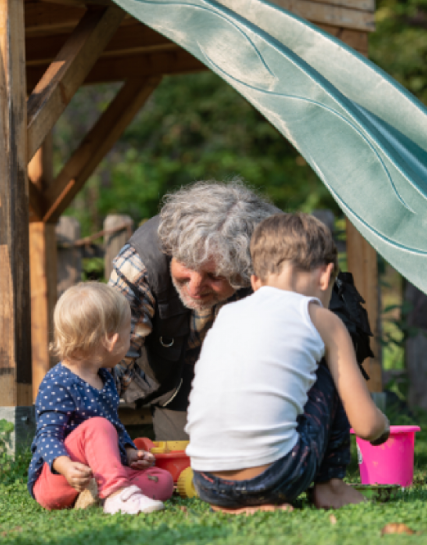 Grandfather kneeling on the grass playing with two grandchildren, outside near a swing set.