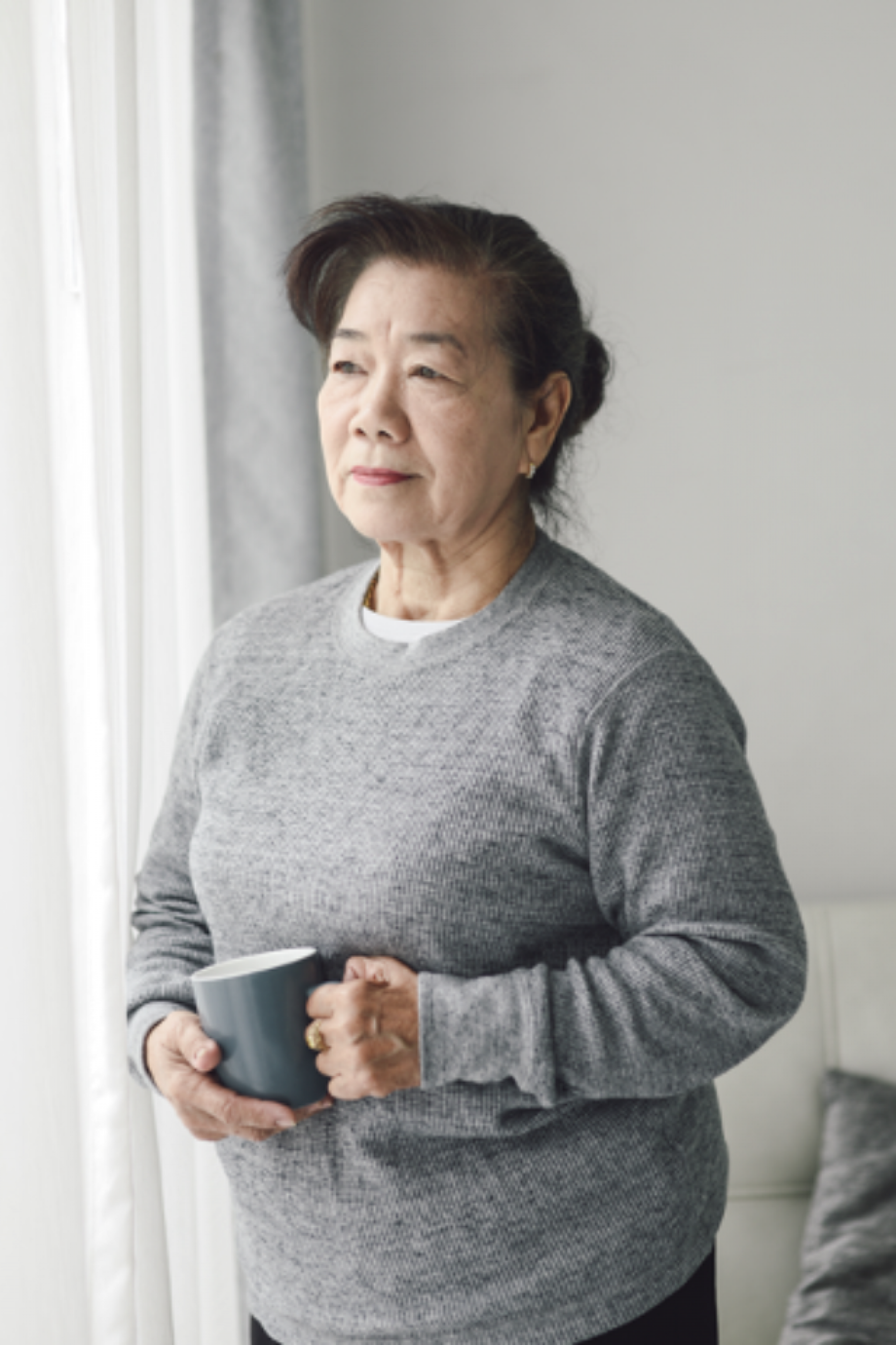 Older adult woman looking out window, holding coffee cup in hand, with thoughtful look on her face.