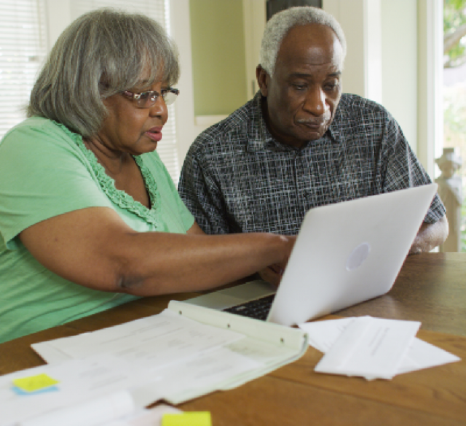 A couple working on documents together, sitting at a kitchen table, also using a laptop.