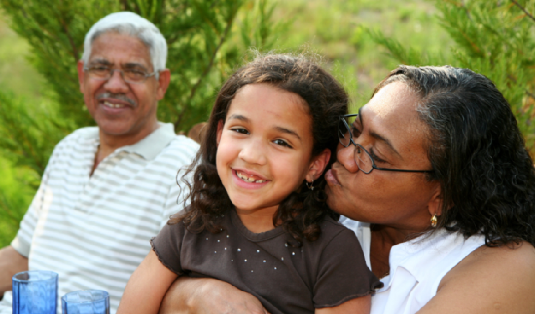 A grandfather, grandmother, and granddaughter sitting together. Grandfather is smiling as grandmother kisses granddaughter on the cheek.