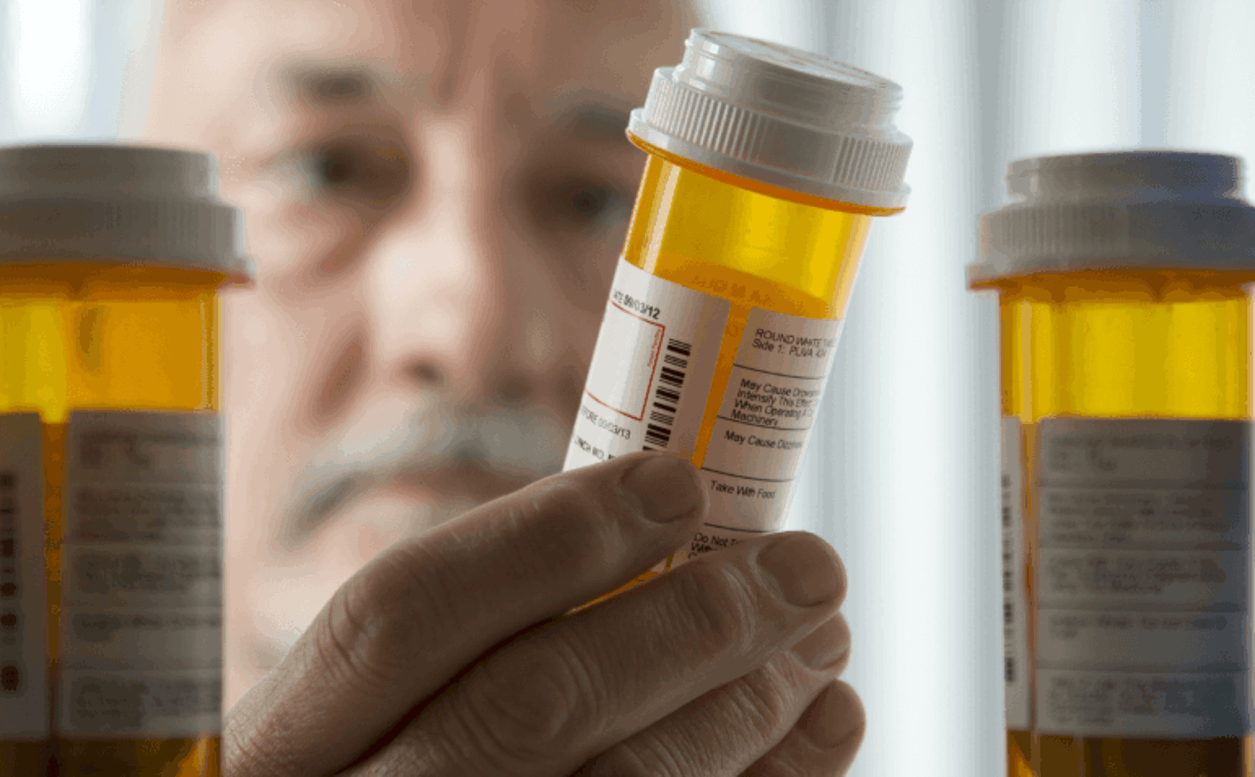 View from inside the medicine cabinet - pill bottles obscure view of older adult looking at one vial of medications.