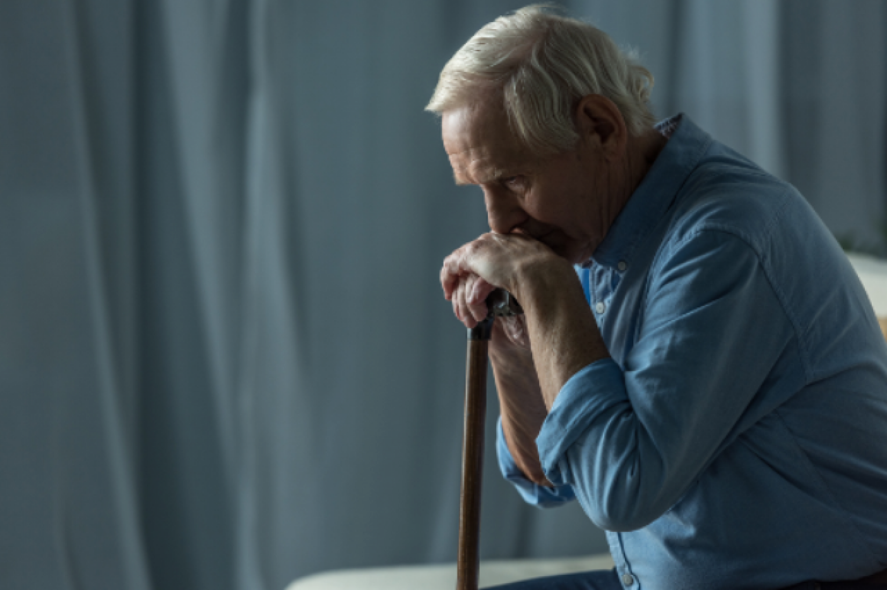 Older adult male with his head resting on his hands, resting on his cane - room is darkened and looks gloomy.